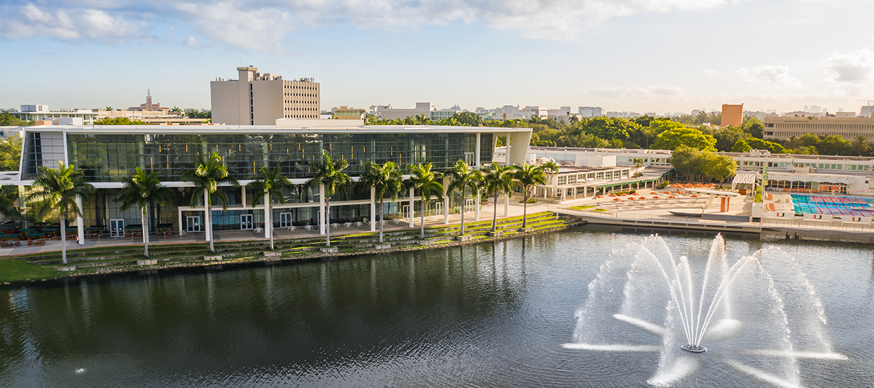 This is an aerial photo of Lake Osceola and the Shalala Student Center at the University of Miami Coral Gables campus. The Coral Gables city scape is in the background, The athletic swimming pool, court yard, and Shalala Student Center are all in the mid-ground. Lake Osceola and a water fountain are in the foreground. 