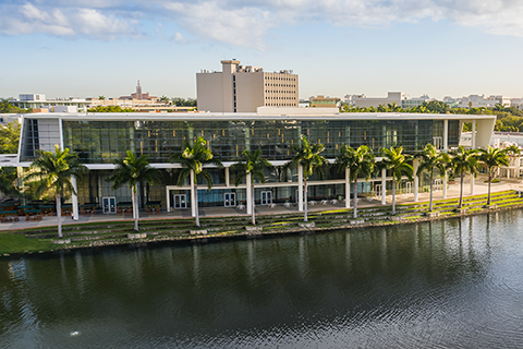 This is an aerial photo of Lake Osceola and the Shalala Student Center at the University of Miami Coral Gables campus. The Coral Gables city scape is in the background, The athletic swimming pool, court yard, and Shalala Student Center are all in the mid-ground. Lake Osceola and a water fountain are in the foreground.