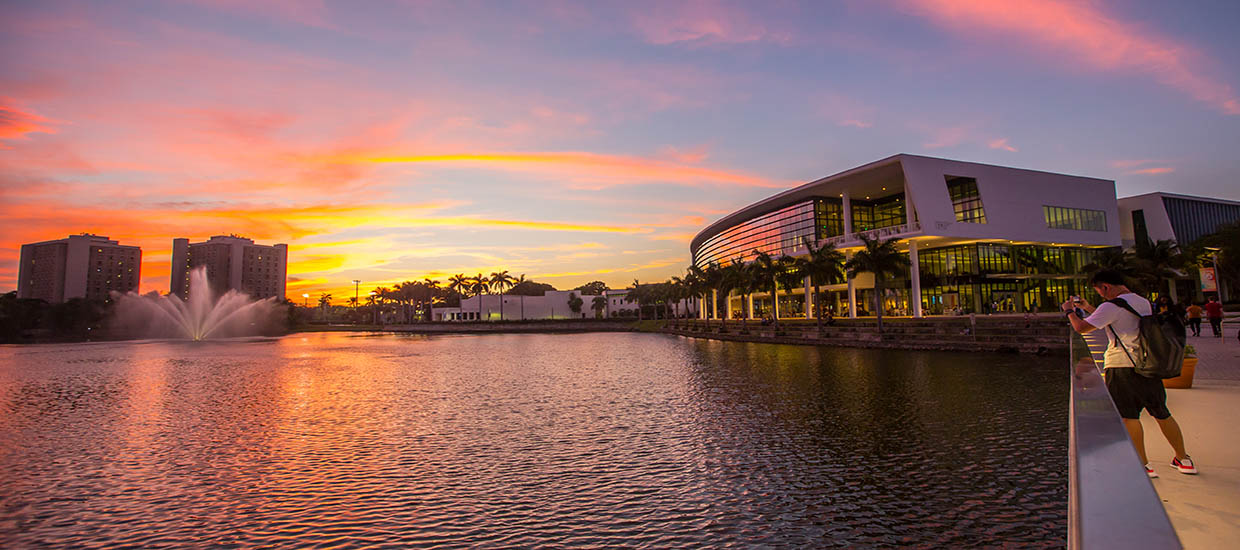 This is a photo of the Shalala Student Center and Lake Osceola at the University of Miami Coral Gables campus. The photo was taken at Sunset and a student is standing on a bridge taking a photo of the sunset over the lake.