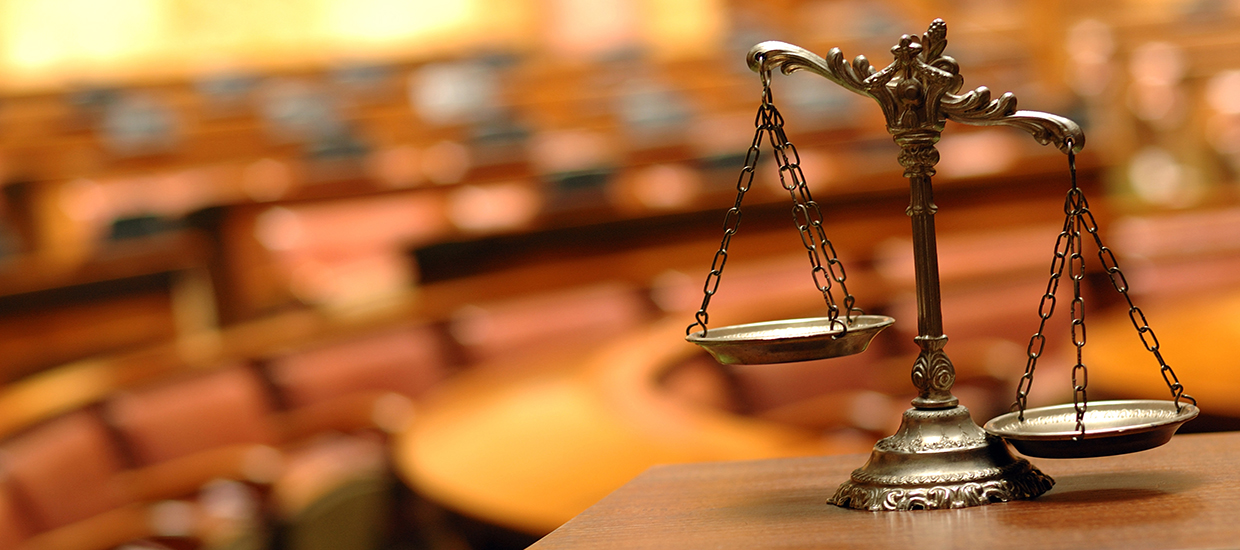 This is a stock photo from Shutterstock. In the foreground are the "scales of justice." The background is blurred. Court room seating is in the background. 