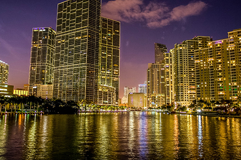 This is a stock photo from Shutterstock. This is an image of the Brickell neighborhood at night. The Biscayne Bay is in the foreground of the image. Brightly lit high-rise buildings are in the background.