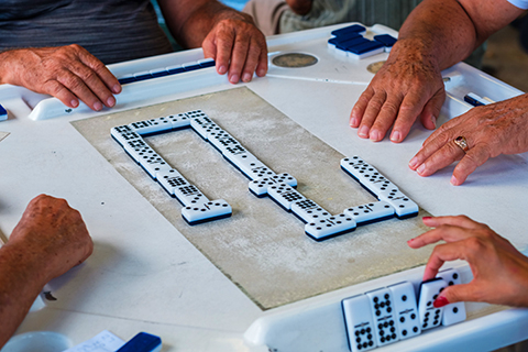 This is a stock photo from Shutterstock. This is an up close image of a Dominos game. Four sets of hands are seen on the table, with a Domino game in play.