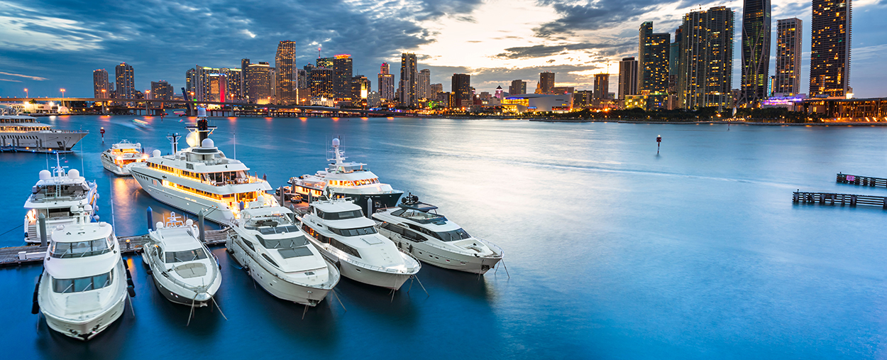 This is a stock photo from Shutterstock. The downtown Miami city scape is in the background. The Biscayne Bay and a dock with several boats is in the foreground. The photo was taken at dusk.