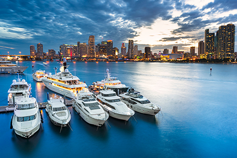 This is a stock photo from Shutterstock. The downtown Miami city scape is in the background. The Biscayne Bay and a dock with several boats is in the foreground. The photo was taken at dusk.
