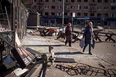 This is a photo of the current events in Ukraine. Two people are walking through a damaged courtyard. Buildings have been destroyed and debris is scattered everywhere. 